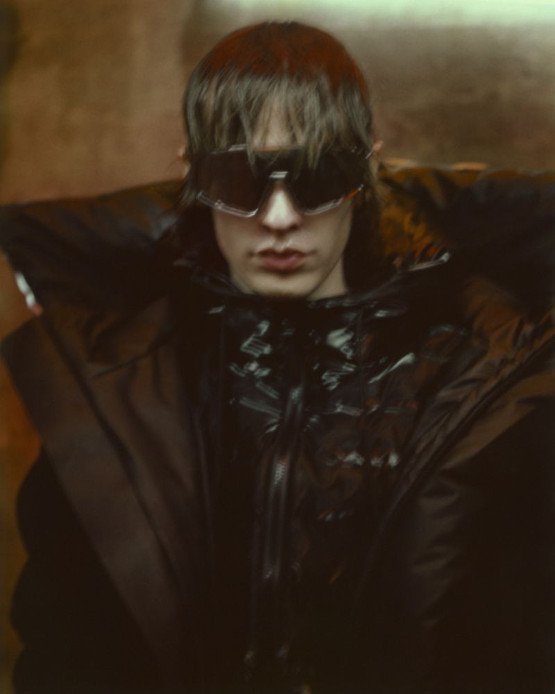 Portrait of a young adult with dark hair wearing futuristic sunglasses and a dark, oversized jacket against an indistinct background.