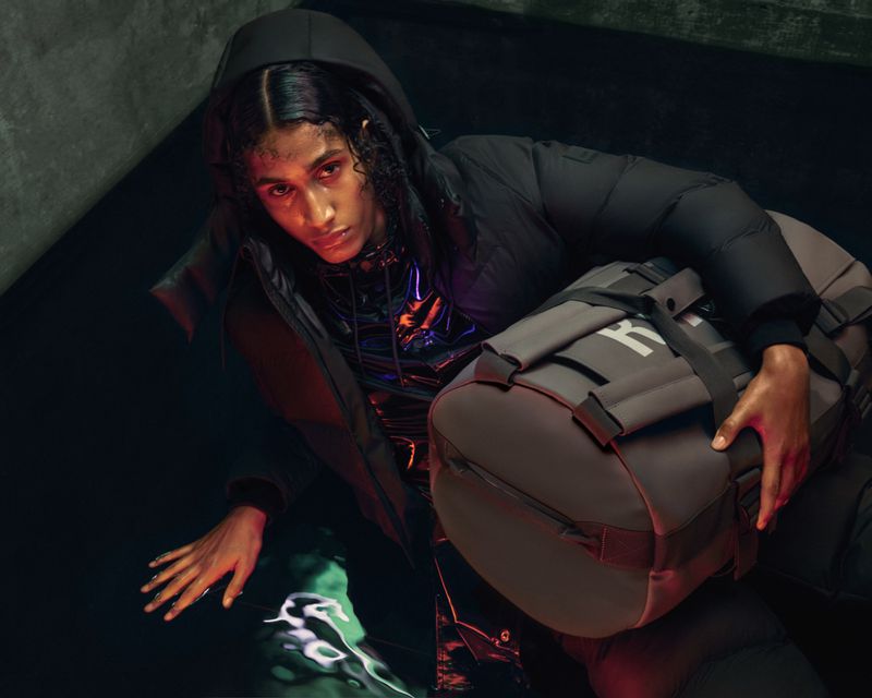Stylish person in hooded puffer jacket holds grey duffel bag in dimly lit setting with reflective pool creating abstract shapes.