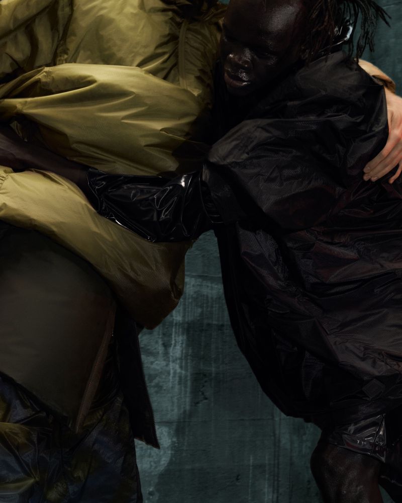 Two people in jackets hug in a low-light fashion shot against a textured, moody wall. A blend of olive and black tones creates an edgy look.