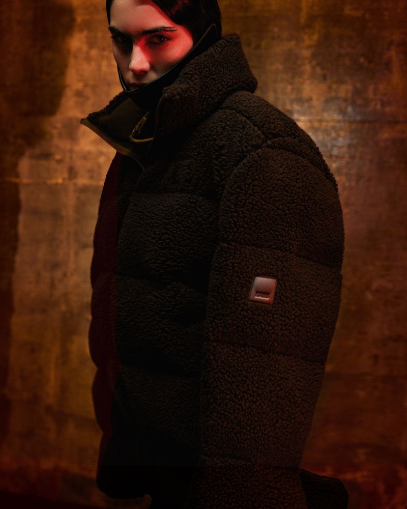Close-up of a young adult woman wearing a dark, puffy coat, illuminated by red light, set against a textured tan background.