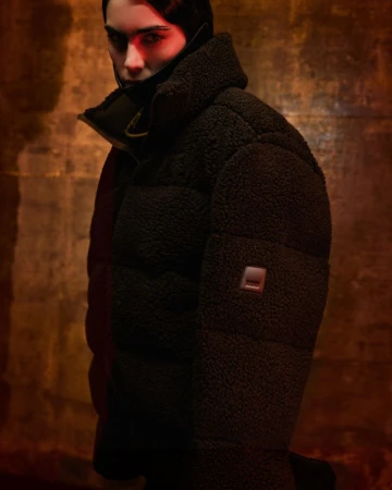 Close-up of a young adult woman wearing a dark, puffy coat, illuminated by red light, set against a textured tan background.
