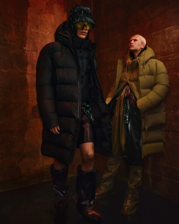 Two fashion models in long puffer coats and boots pose in a photo studio with a textured red-brown wall as the backdrop.
