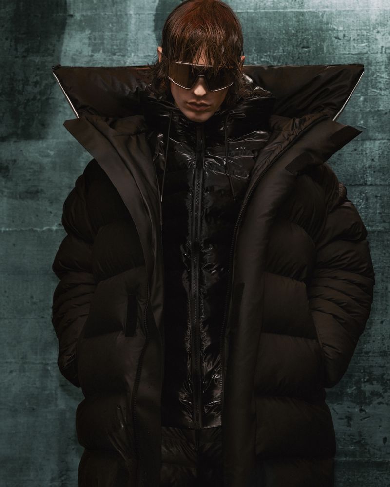 Fashion portrait of a person wearing a black puffer coat and silver sunglasses, with a green textured background, eye-level angle.
