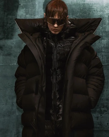 Fashion portrait of a person wearing a black puffer coat and silver sunglasses, with a green textured background, eye-level angle.