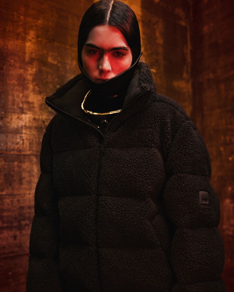 Model wearing an oversized black textured fleece puffer jacket with a structured high collar in dramatic warm studio lighting.