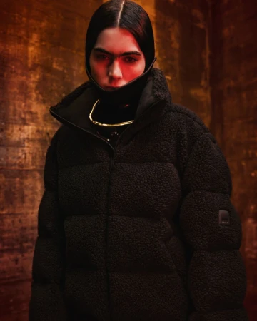 Model wearing an oversized black textured fleece puffer jacket with a structured high collar in dramatic warm studio lighting.