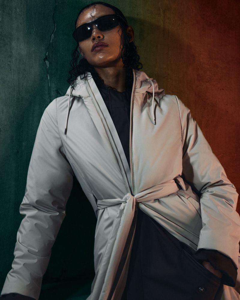 Person in a beige raincoat and sunglasses poses against a distressed teal and orange wall in a fashion or beauty editorial shot.