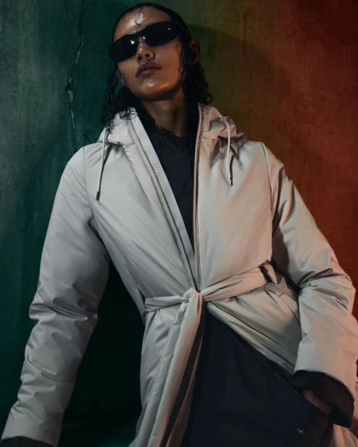 Person in a beige raincoat and sunglasses poses against a distressed teal and orange wall in a fashion or beauty editorial shot.