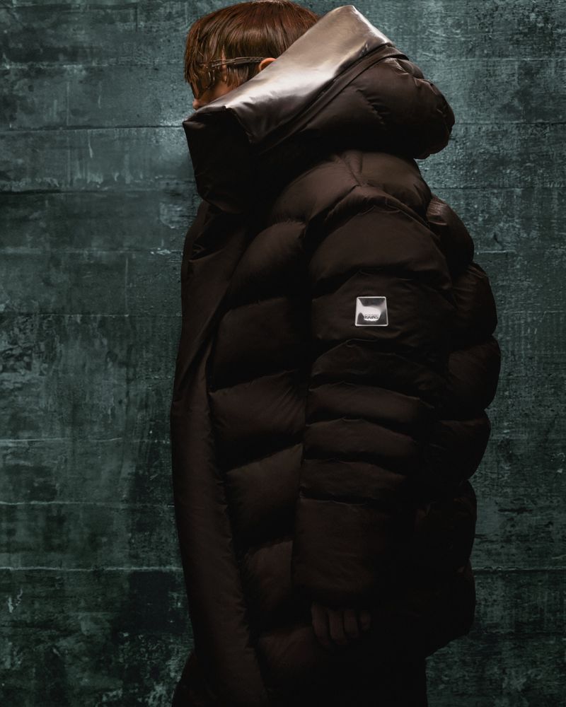 An adult wears a dark brown puffy down coat with a high collar. The shot is a studio setting against a dark grey textured background.