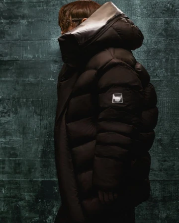 An adult wears a dark brown puffy down coat with a high collar. The shot is a studio setting against a dark grey textured background.