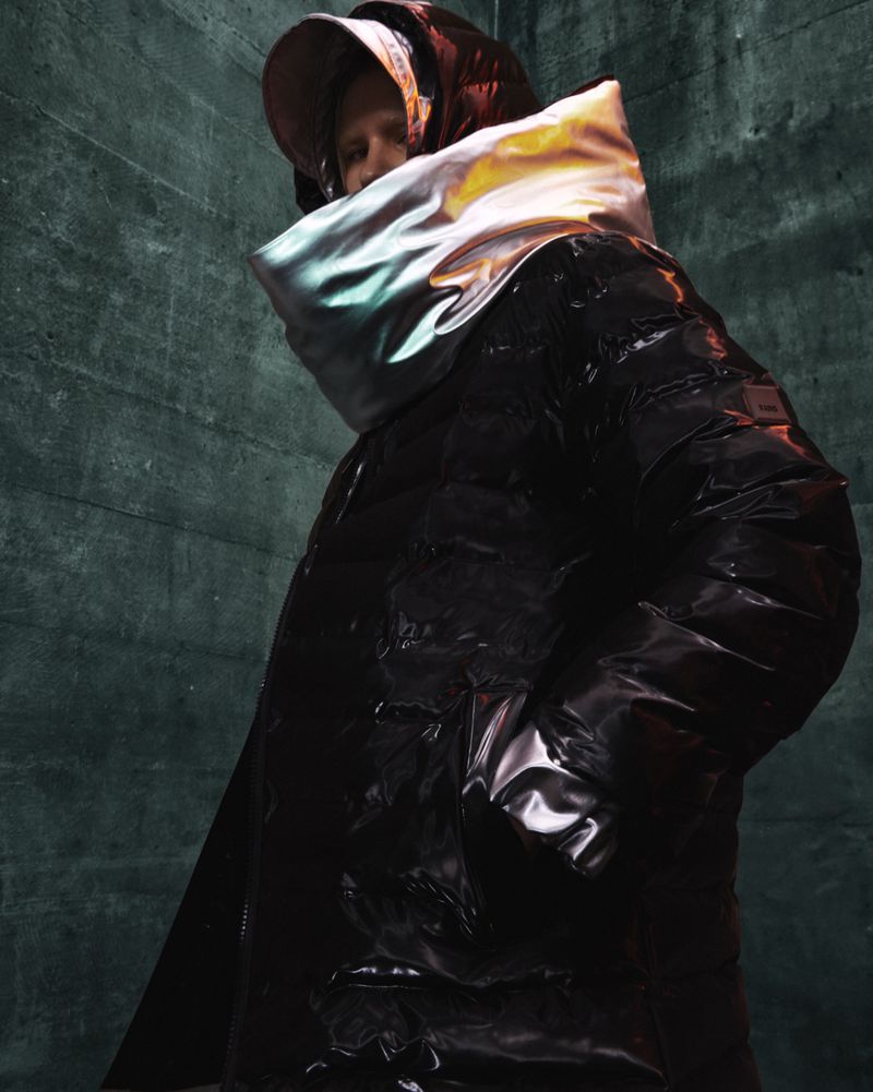 Fashion shot featuring an androgynous person in a black puffer jacket with a high collar, standing against a textured wall.