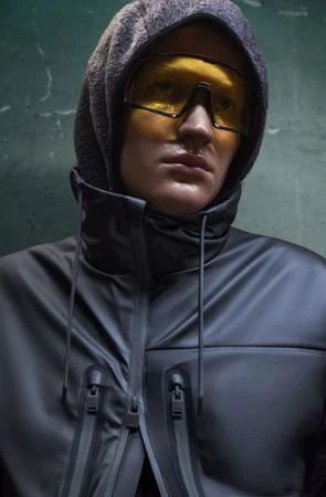 Close-up of a hooded person with yellow sunglasses wearing a dark, rubbery jacket with visible zippers against a textured green wall.