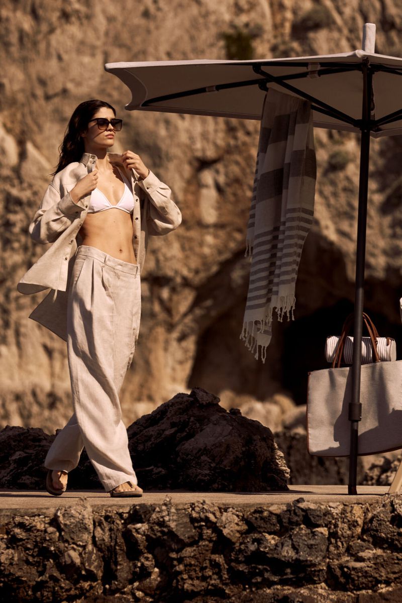 A woman in a white bikini top and tan linen pants and button-down shirt stands on a stone patio with a beach umbrella.