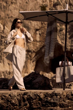 A woman in a white bikini top and tan linen pants and button-down shirt stands on a stone patio with a beach umbrella.