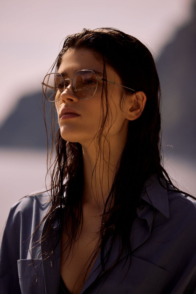 Portrait of a brunette woman with long, wet hair wearing sunglasses and a blue shirt against a blurred outdoor backdrop.