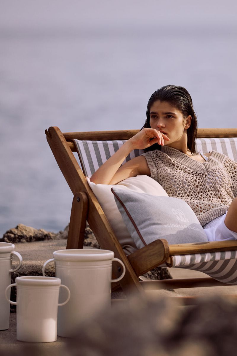 Woman lounges on beach chair with cushion and mugs, enjoying serene outdoor setting with the sea in the distance on a cloudy day.