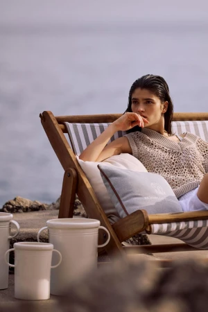 Woman lounges on beach chair with cushion and mugs, enjoying serene outdoor setting with the sea in the distance on a cloudy day.