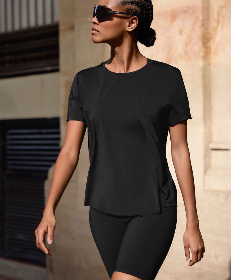 Athletic woman walks in black sportswear and sunglasses, confidently exercising outdoors in urban setting, promoting fitness lifestyle.