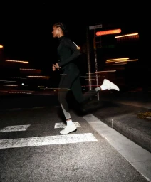 Woman running across an asphalt crosswalk at night in black sportswear and white sneakers.
