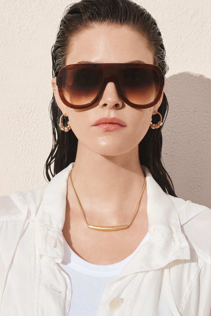 Close-up studio portrait of a woman with slicked-back hair wearing large sunglasses, gold hoop earrings, a gold necklace, and a white shirt.