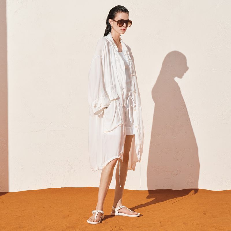 A woman models in white oversized shirt, shorts, and sandals against a bright wall. The image showcases fashion and lifestyle in simple setting.