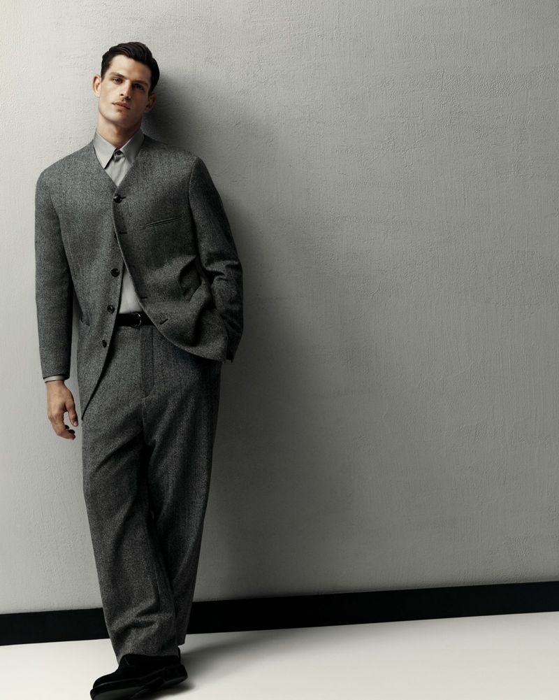 Man posing, leaning against a grey wall, wearing a grey suit, dress shirt, black belt, and black shoes in a studio setting.