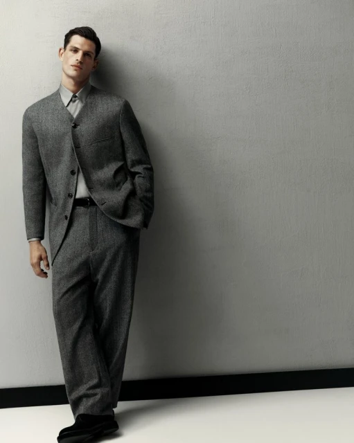 Man posing, leaning against a grey wall, wearing a grey suit, dress shirt, black belt, and black shoes in a studio setting.