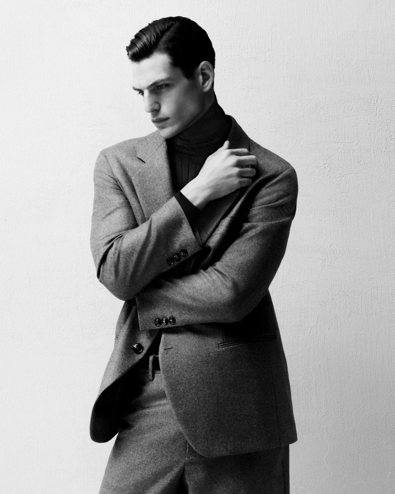 Monochrome portrait of a man in a suit with his arms crossed, wearing a turtleneck, standing indoors against a textured white wall.