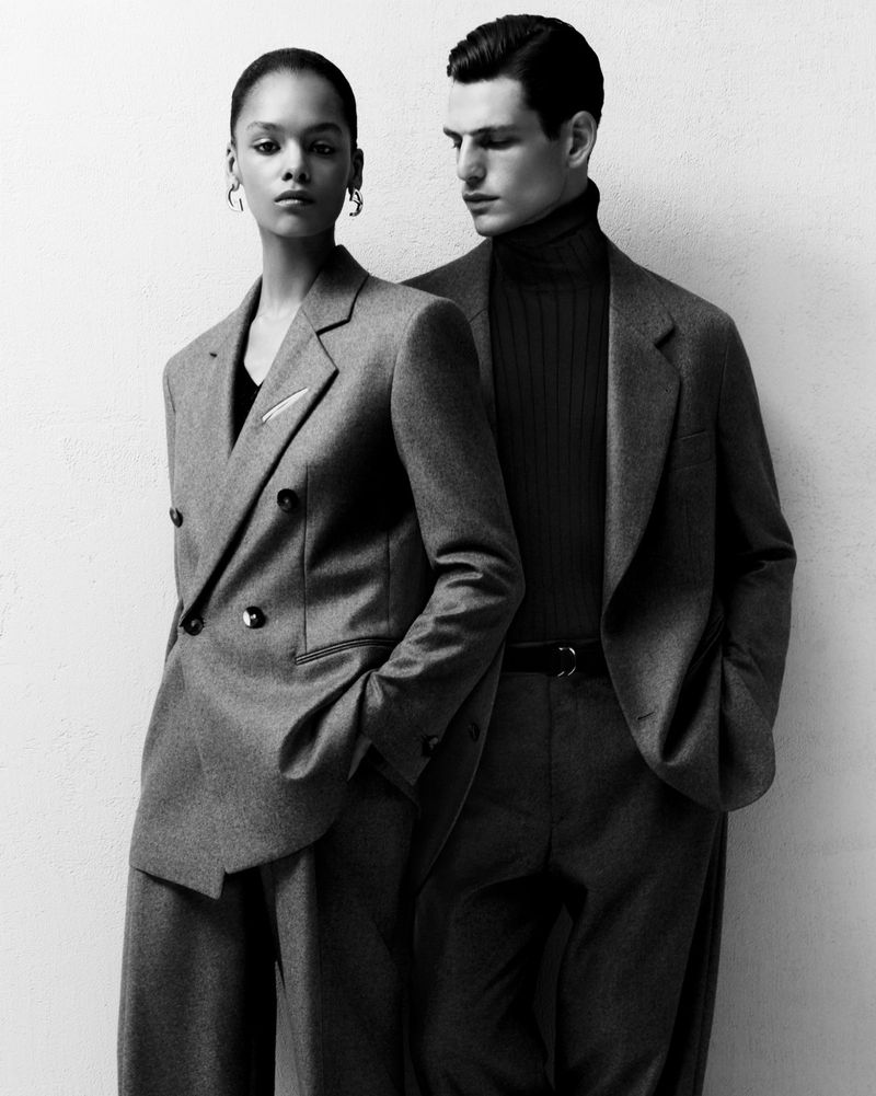 Black and white studio shot of a Black female model in a gray pantsuit and a white male model in a gray suit and turtleneck.