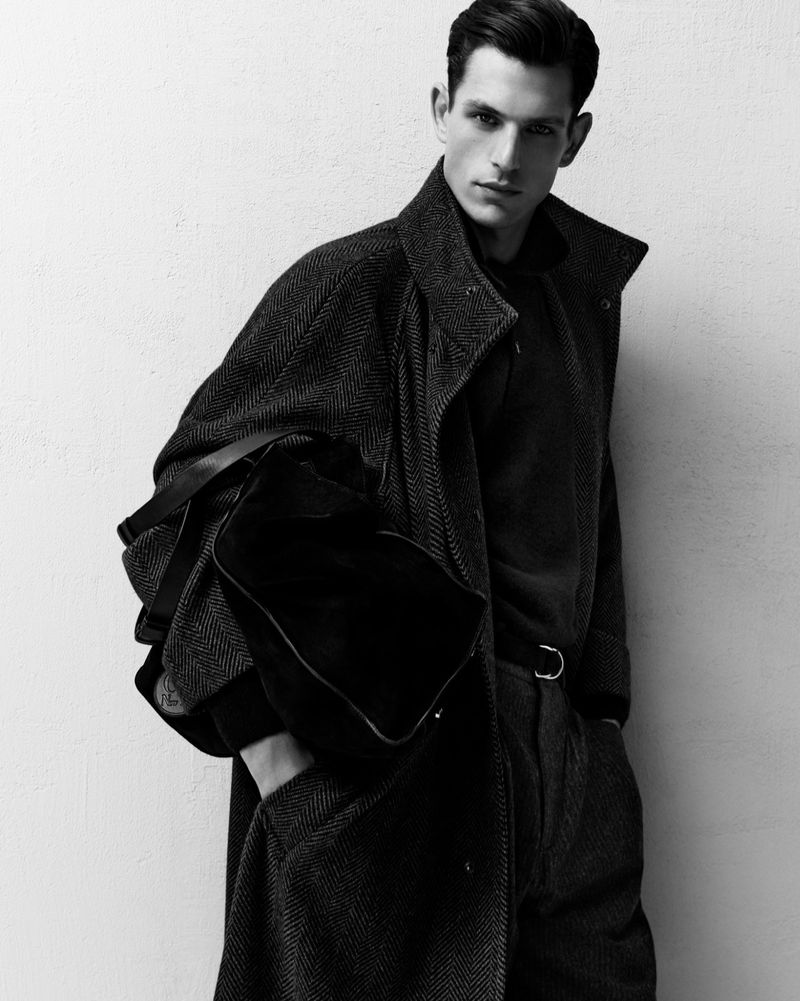 Monochrome portrait of a male model wearing a herringbone coat holding a black duffel bag against a plain wall