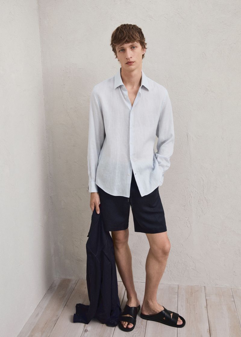 A male fashion model poses in a studio wearing a button-down shirt, shorts, and sandals, holding a jacket, in a casual pose.