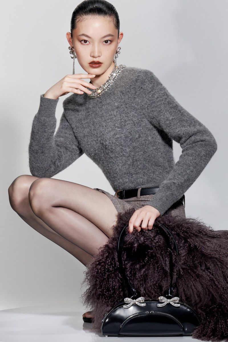 Asian woman crouching indoors in a gray sweater, black tights, and a black handbag with decorative bows. Fashion-forward studio shot.