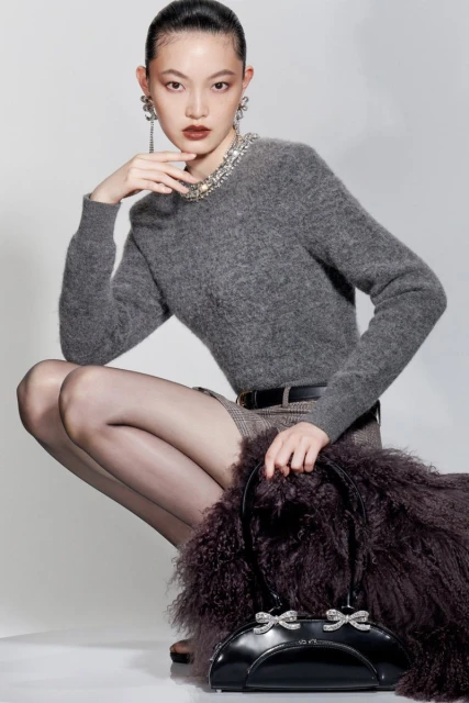 Asian woman crouching indoors in a gray sweater, black tights, and a black handbag with decorative bows. Fashion-forward studio shot.