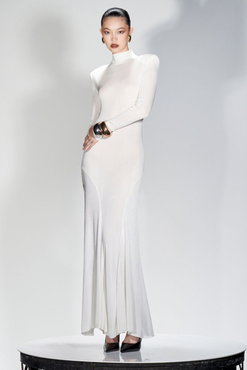 An Asian woman is posing in a full-length studio shot wearing a white, form-fitting gown, high heels, and gold bracelet against a bright background.