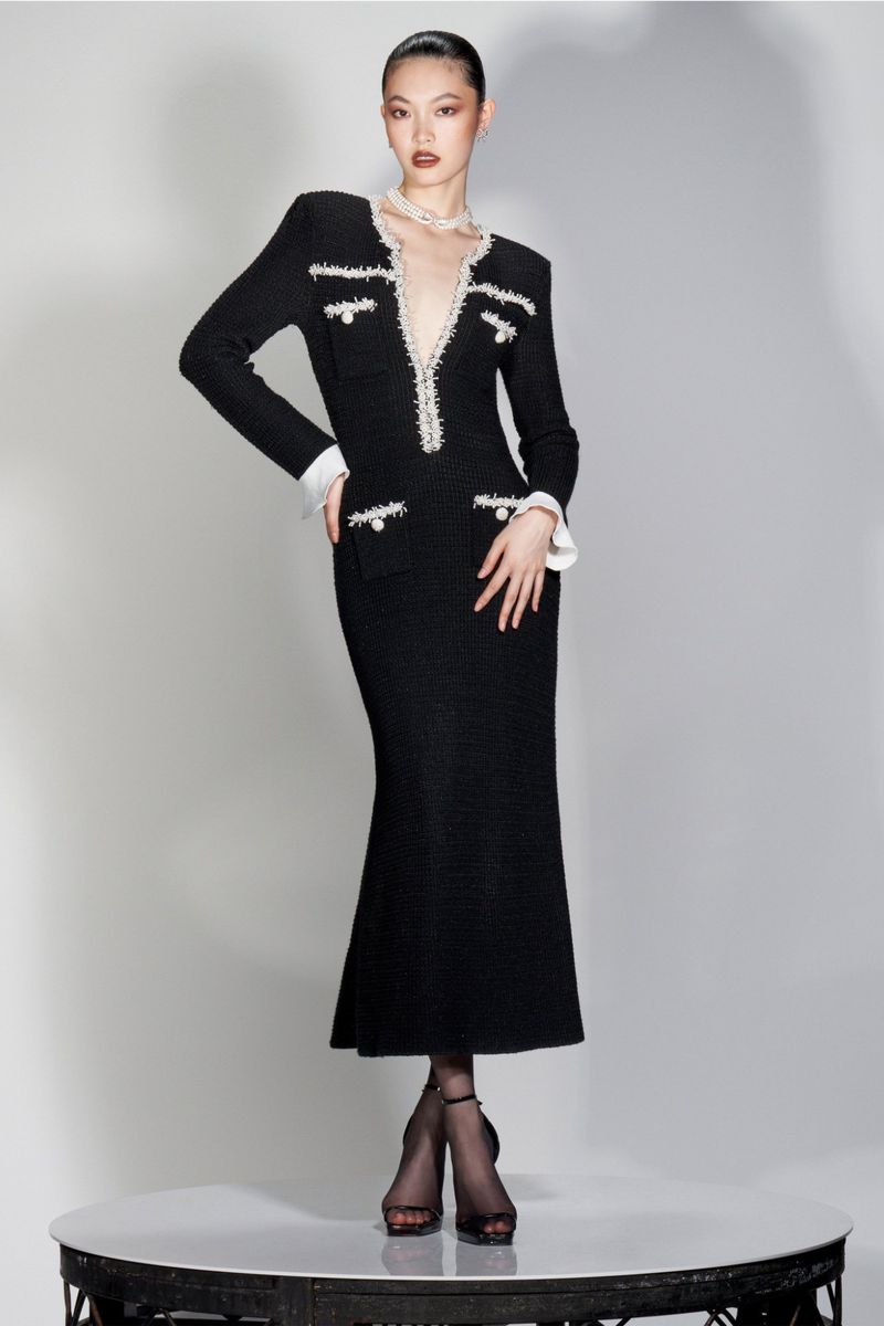 Elegant Asian woman poses in a stylish black tweed midi dress with pearl accents and sheer black tights on a round platform in a studio.