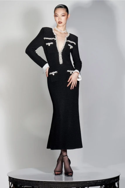 Elegant Asian woman poses in a stylish black tweed midi dress with pearl accents and sheer black tights on a round platform in a studio.