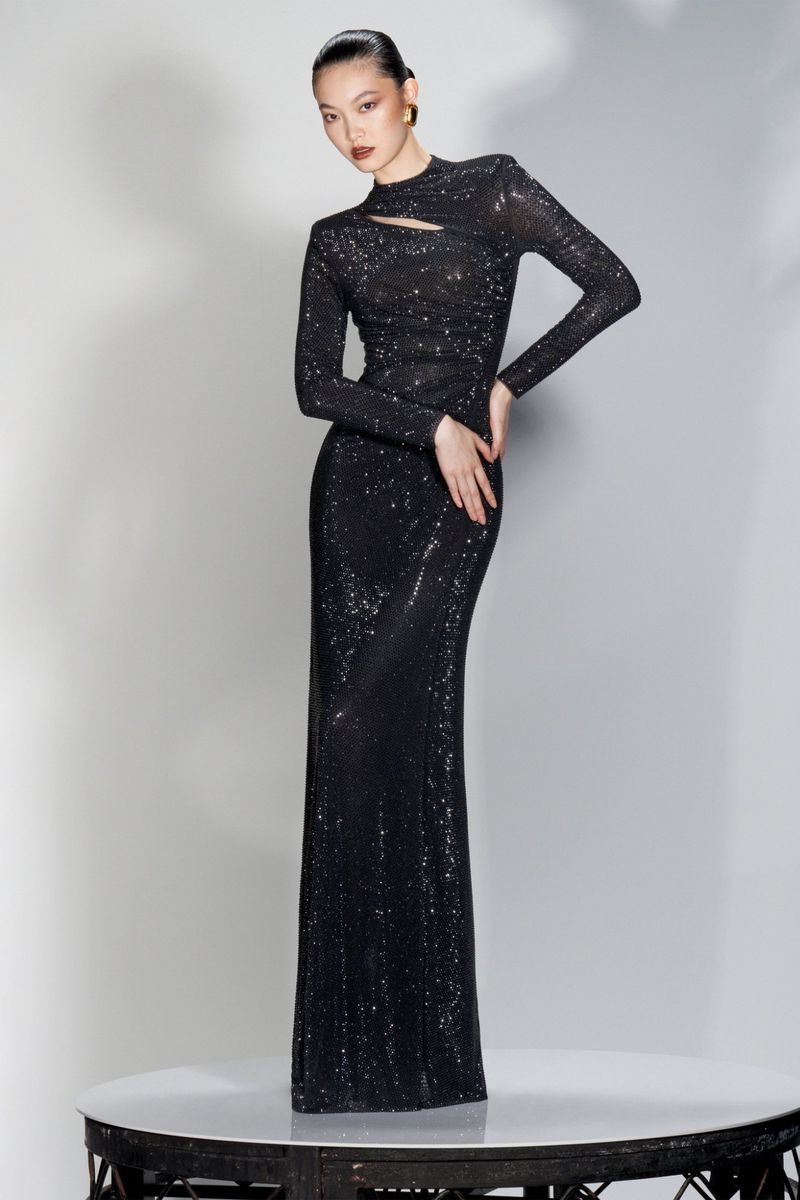 Elegant woman in long-sleeved, floor-length, sparkly black gown stands on a round table against a neutral background in a studio shot.
