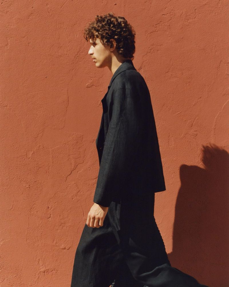 Fashionable male model in dark suit walks in front of coral textured wall in fashion editorial photography, outdoor setting, daytime.