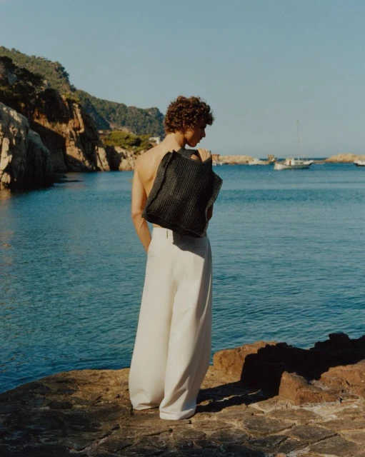 A person stands on a rocky shore carrying a tote bag, with a calm blue sea, cliffs, and boats in the background on a bright day.