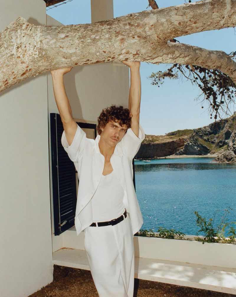 Young man in casual white clothing leans against a tree branch overlooking ocean and rocky coastline on a sunny day. 