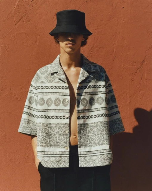 Adult man wearing patterned button-down shirt and black bucket hat standing against an orange wall in an indoor setting.