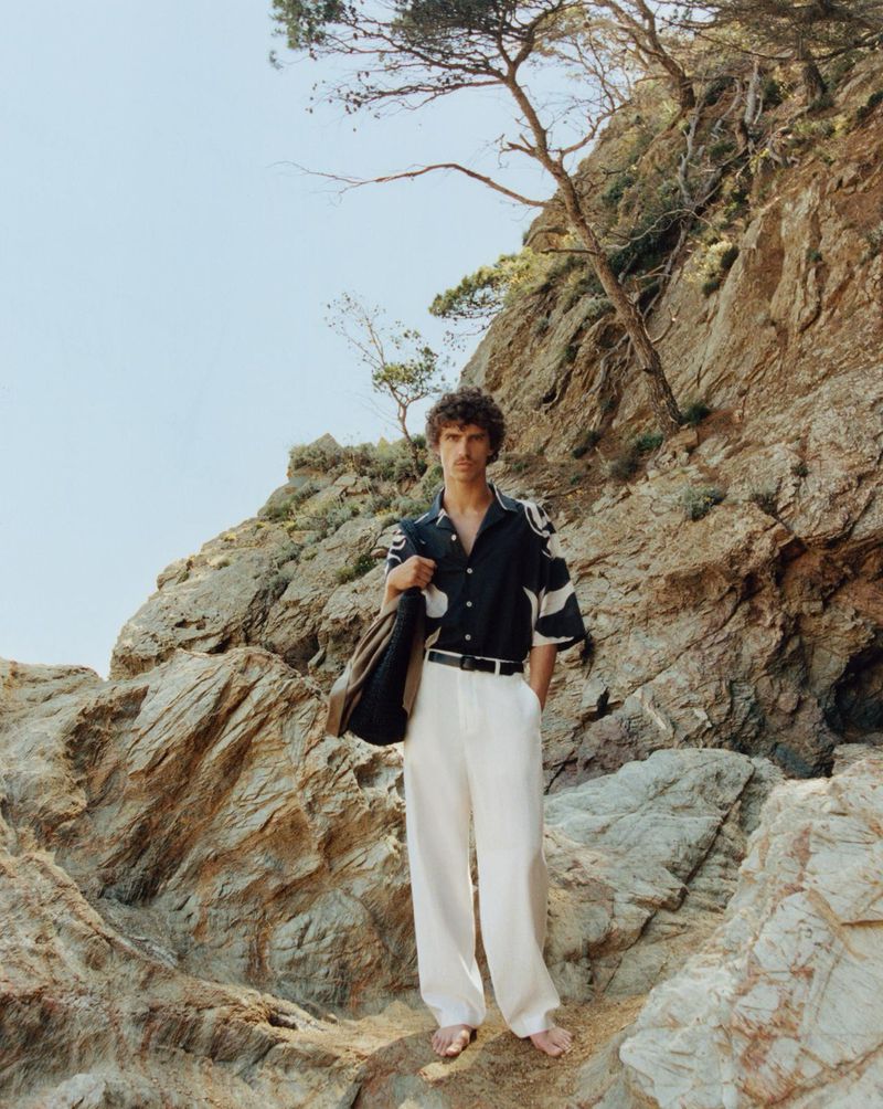 Young man in patterned short-sleeve shirt and white pants stands on rocks near the coast with a shoulder bag in daylight.