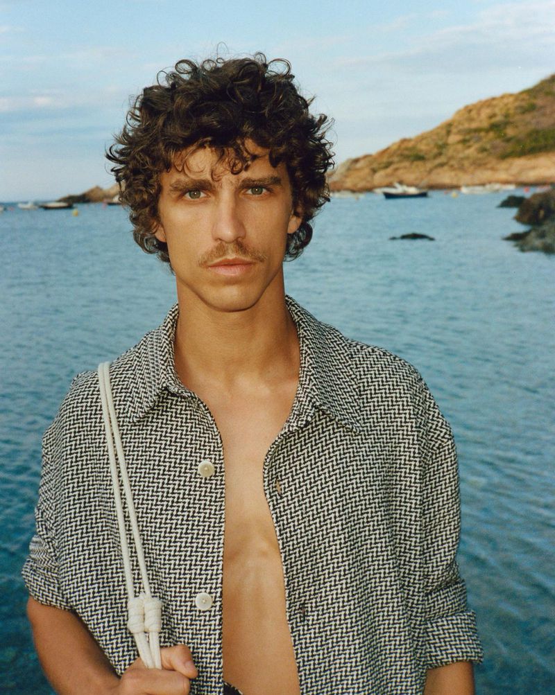 An adult male poses outdoors wearing an open button-down shirt with a pattern, near the ocean, with boats and rocks in the background.