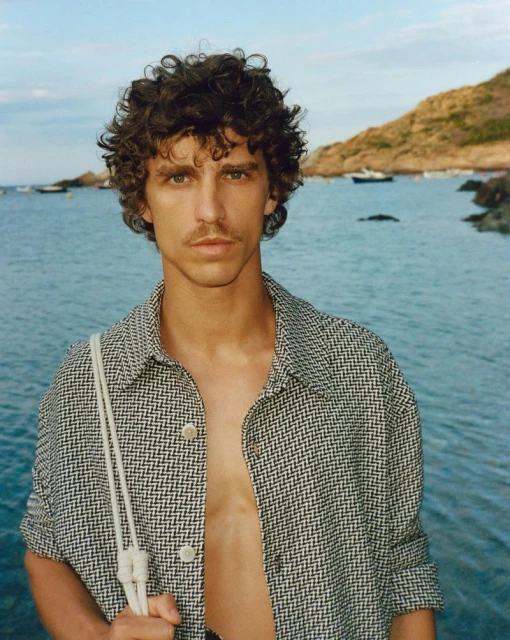 An adult male poses outdoors wearing an open button-down shirt with a pattern, near the ocean, with boats and rocks in the background.