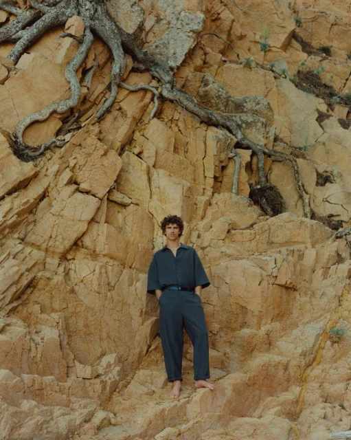Adult male in blue jumpsuit poses barefoot against a rocky cliff face with tree roots overhead in an outdoor setting on a sunny day.