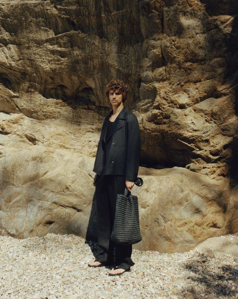 Adult man in dark jacket and pants stands with a woven tote bag in an outdoor setting with a large rock face backdrop.
