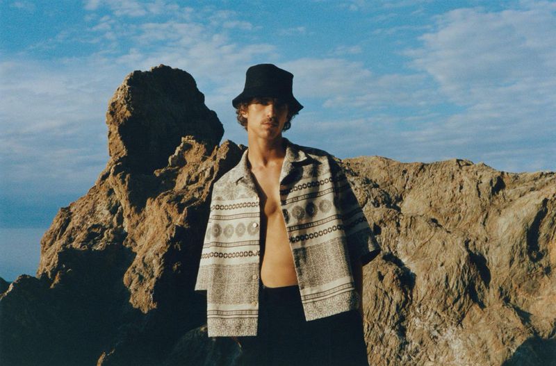 Man poses in patterned shirt and black bucket hat against a rocky landscape under a partly cloudy sky on a sunny day.