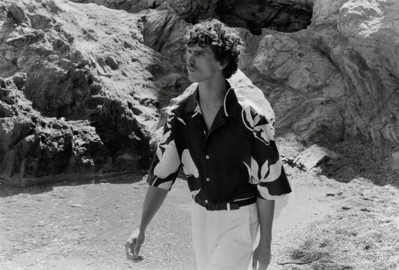 Model Grzegorz Chanko wearing a floral print short-sleeve shirt and light trousers along a rocky coastal shoreline in black and white.