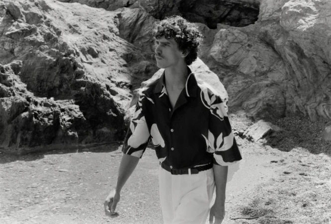 Model Grzegorz Chanko wearing a floral print short-sleeve shirt and light trousers along a rocky coastal shoreline in black and white.