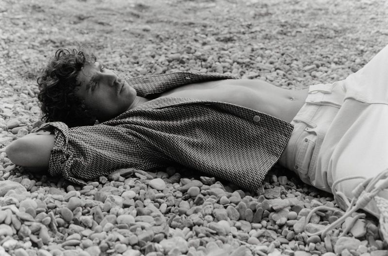 Black and white portrait of a curly-haired male model lying on a rocky beach, wearing an unbuttoned jacket and light pants.